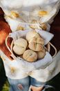 From above of crop faceless female baker in apron and torn jeans with wicker bowl of soft buns with cross cuts on tops standing in sunlight