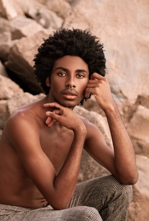 Young trendy African American male with naked torso and piercing in nose touching face while looking at camera and sitting on stones