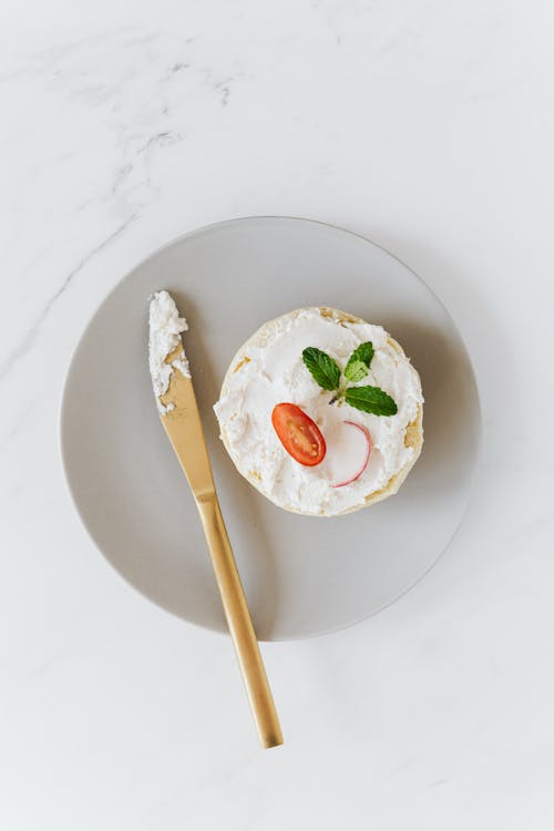 Top view of plate with bun with cottage cheese spread and tomato with radish decorated with small mint sprig on round platter with gold plated knife