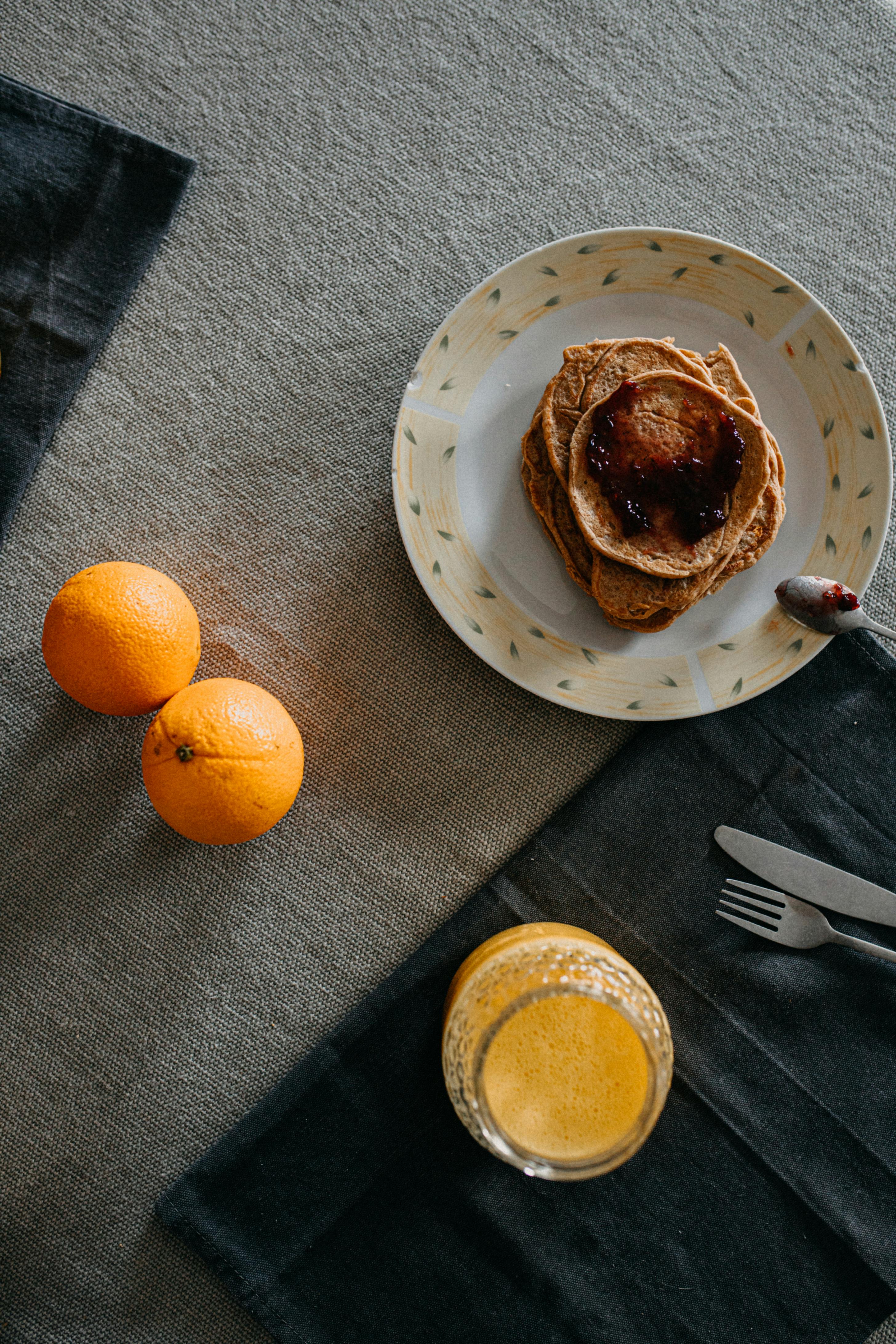 Overhead view of pancakes with jam, oranges, and orange juice on a rustic tablecloth.