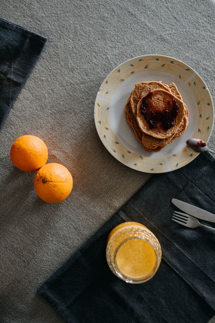 Overhead view of pancakes with jam, oranges, and orange juice on a rustic tablecloth.
