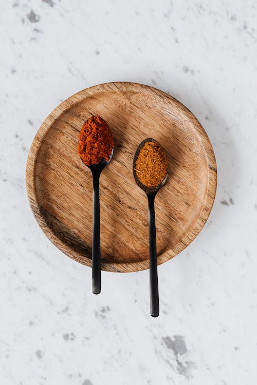 Top view of round wooden plate with spoons filled with ground paprika and mix of peppers on marble table