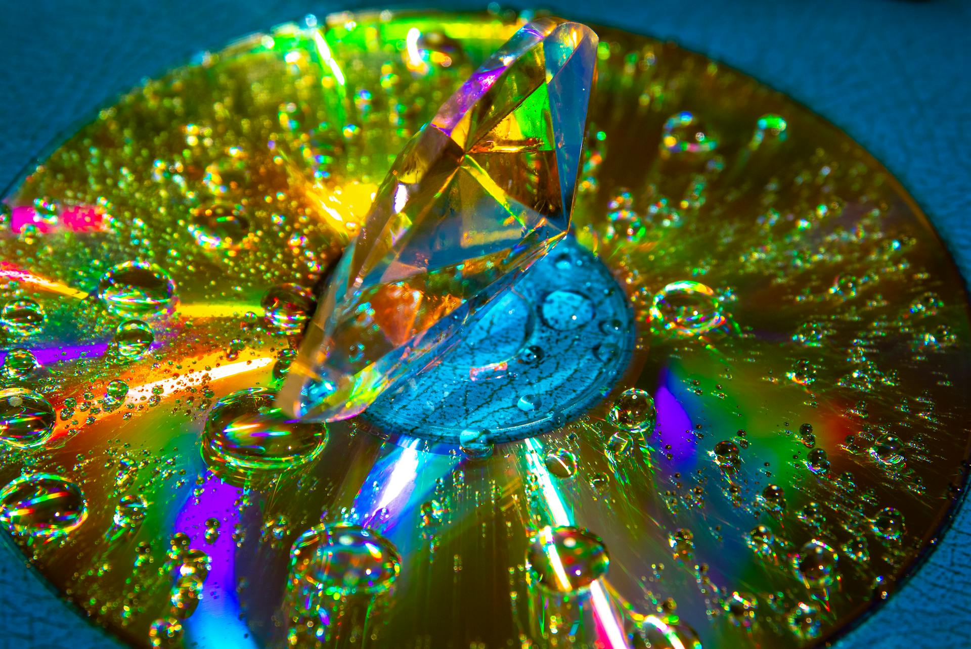 A colorful prism diamond resting on a CD reflecting rainbow colors with water droplets creating a vivid light display.