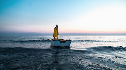 Back view of anonymous male in aged boat admiring rippled ocean with horizon under sky at sundown