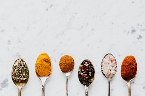 Top view of assorted aromatic condiments arranged together in metal spoons on marble surface used for delicious gourmet meal cooking