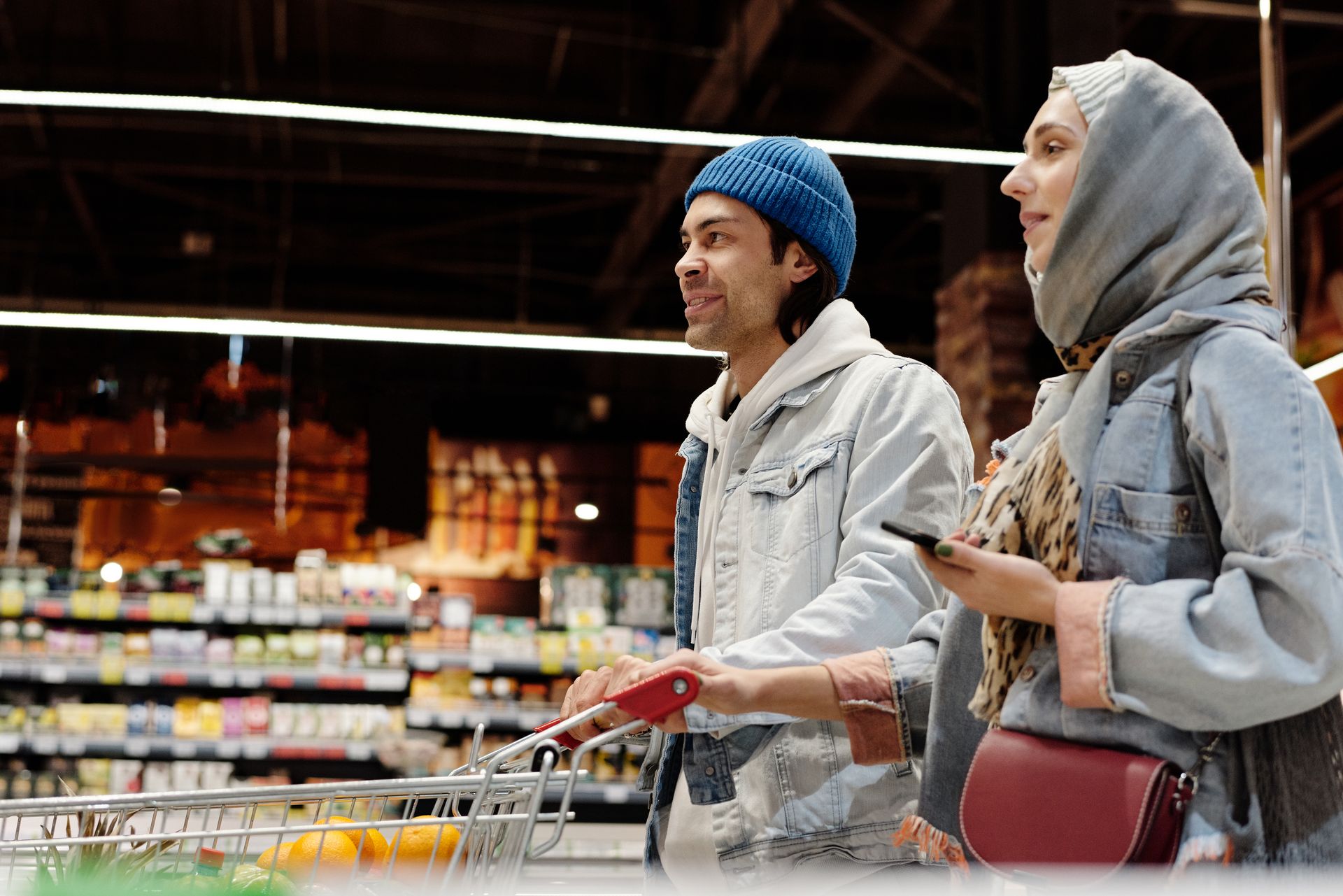 Middle Eastern couple shopping, smiling in supermarket aisle with a cart and smartphone.