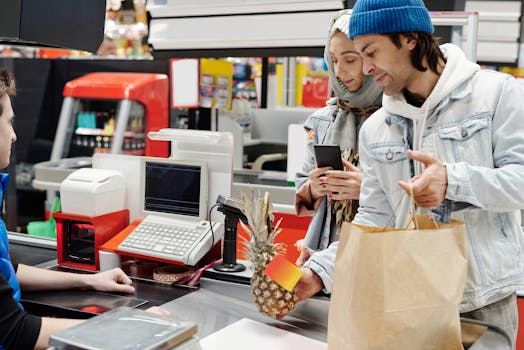 A couple shopping at a supermarket checkout, holding a pineapple and paper bag.
