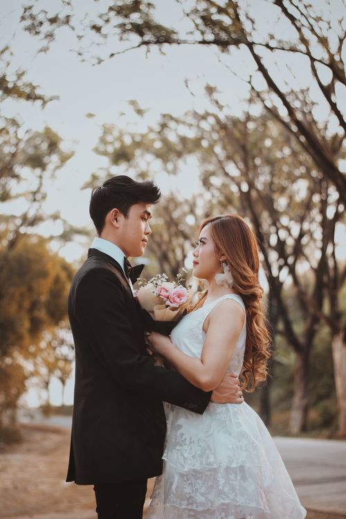 Free Side view of young ethnic male in formal suit embracing charming partner in wedding dress while looking at each other in park Stock Photo