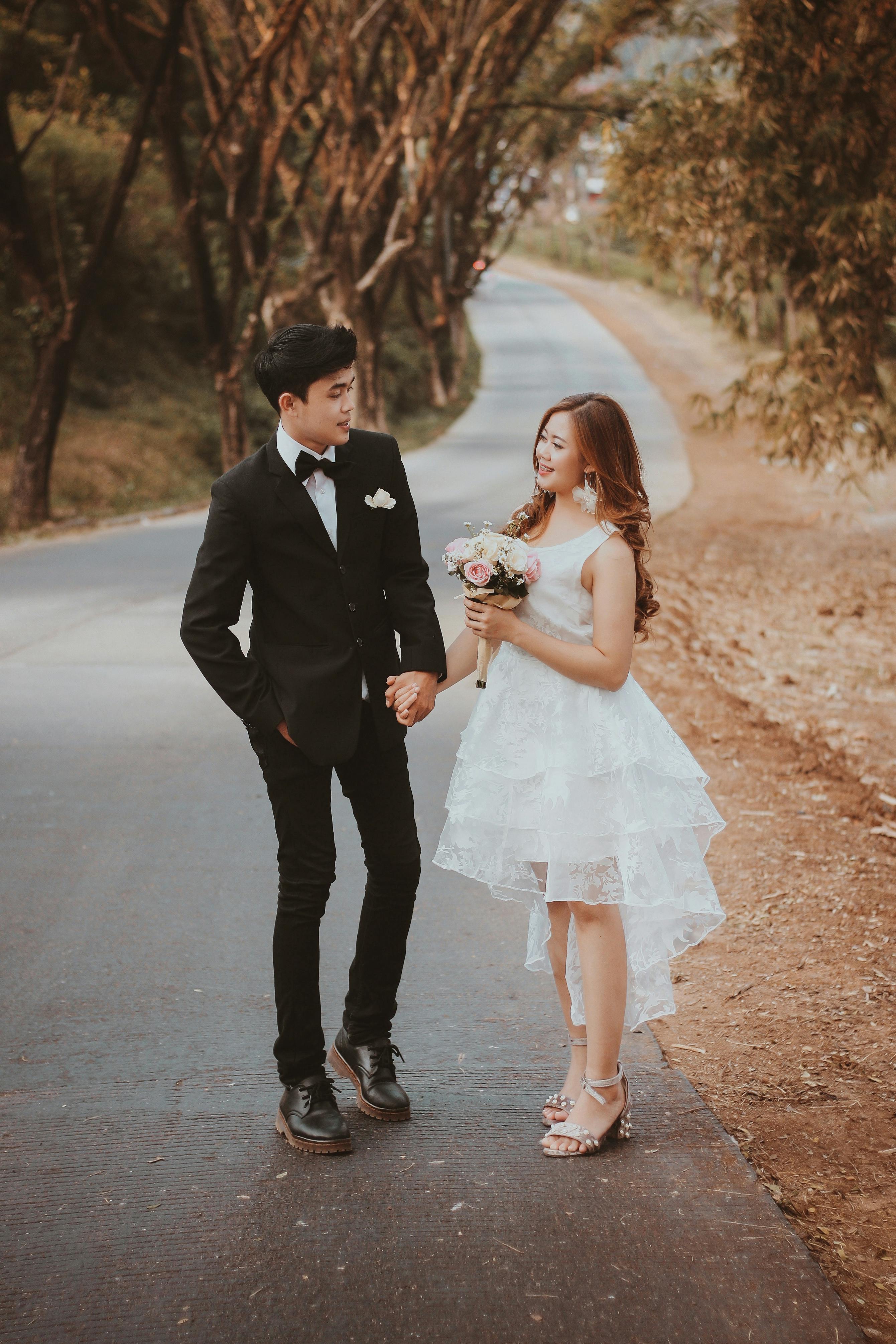 From above of elegant ethnic groom in suit with hand in pocket holding by hand cheerful beloved in white dress with bridal bouquet while looking at each other