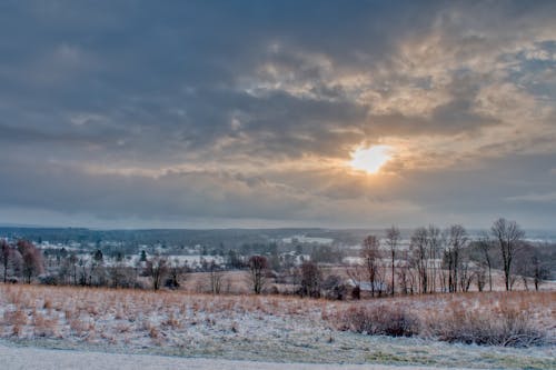 Scenery view of glowing sun in cloudy sky over snowy land with dry trees and grass at sunset