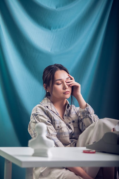 Free Thoughtful young female in casual clothes with closed eyes sitting at white table against blue drapery Stock Photo