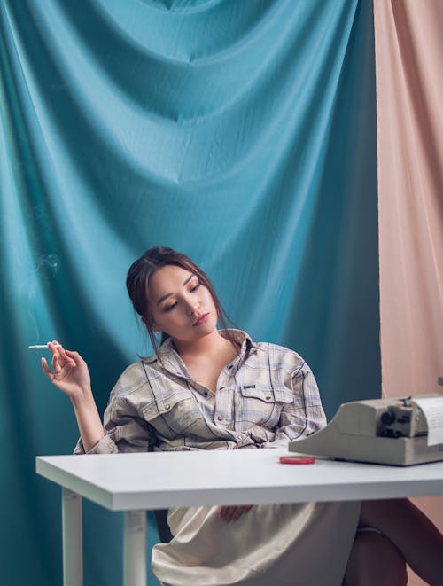 Pensive young female in casual wear sitting at white table near blue and pink drapery while smoking cigarette