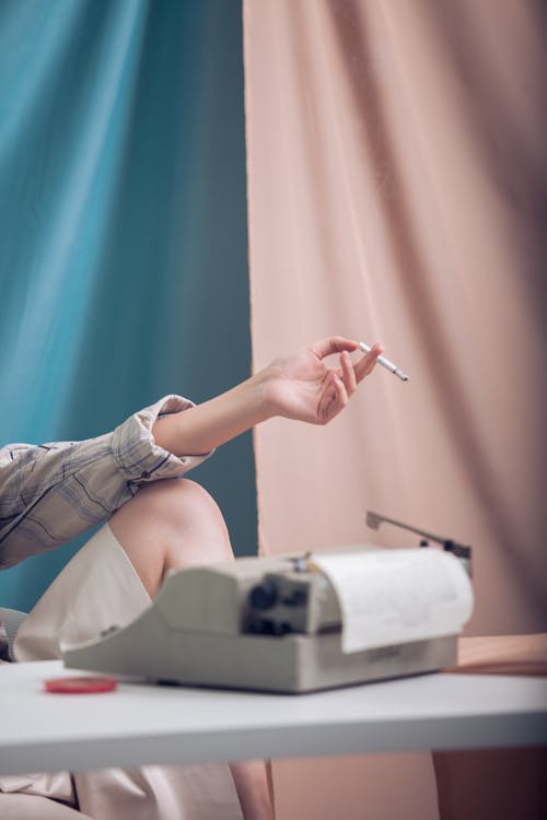 Unrecognizable female sitting at white table with typewriter near blue and pink curtain while holding cigarette in hand