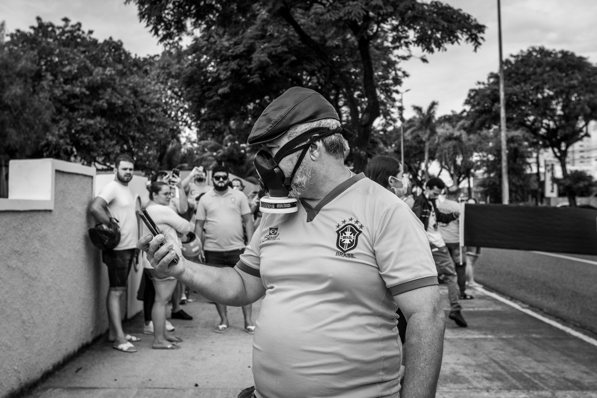 Black and white photo of a man wearing a gas mask using a phone at an outdoor event.