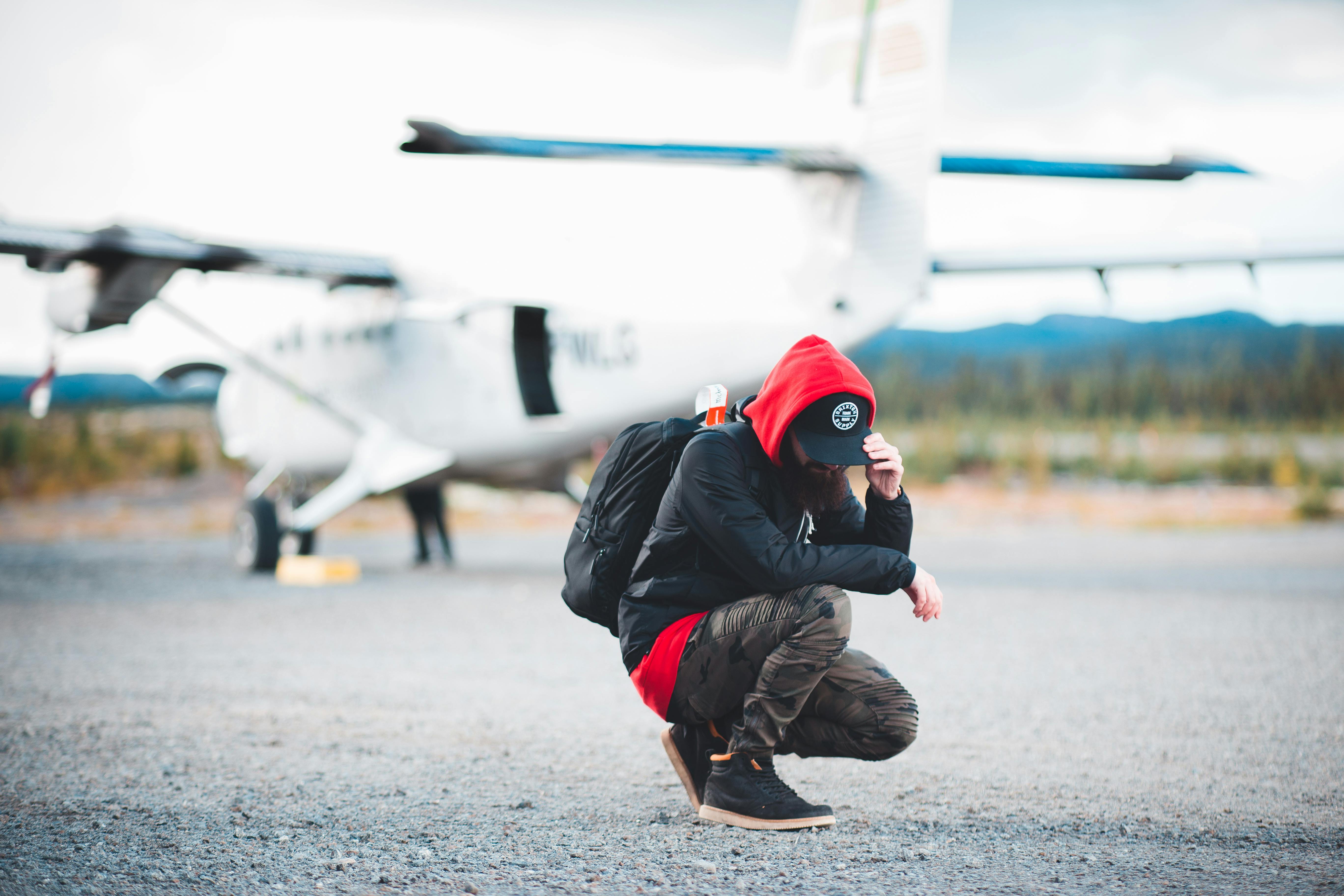 Full length unrecognizable male backpacker wearing casual outfit hunkering down against blurred modern light plane and touching cap peak before flight