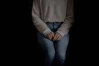 Crop anonymous female in jeans and white sweater sitting coyly with hands on laps in dark studio