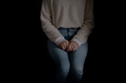Crop anonymous female in jeans and white sweater sitting coyly with hands on laps in dark studio