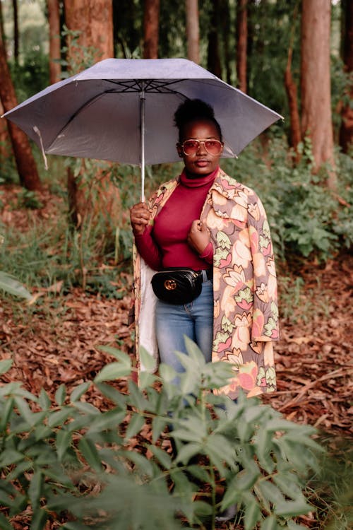 Attractive African American female wearing trendy outfit and coat standing under umbrella amidst green lush trees in forest and looking away calmly