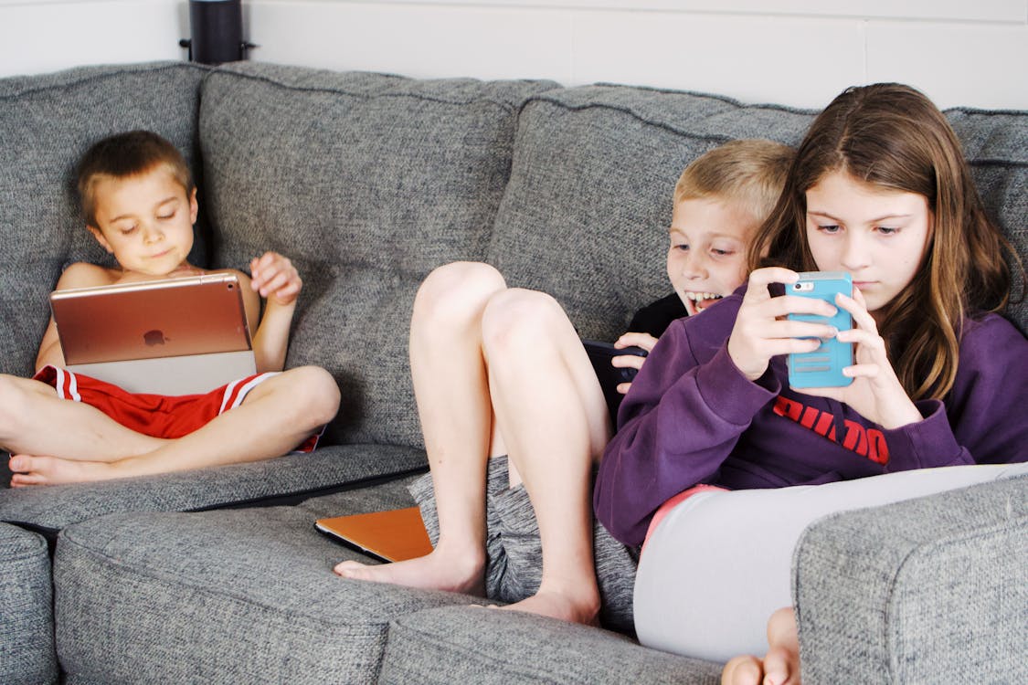 Free Positive barefoot children in casual wear resting together on cozy couch and browsing tablets and smartphones Stock Photo