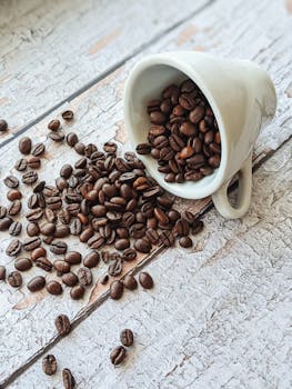 A ceramic cup spilling fresh roasted coffee beans onto a rustic wooden table, creating a minimalistic look.
