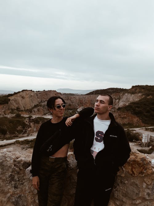 Content man resting arm on friends shoulder while standing together in mountainous terrain during trip