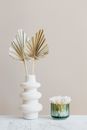 Composition of white ceramic vase with artificial leaves and green glass reusable container for cotton buds placed on white marble shelf against beige wall