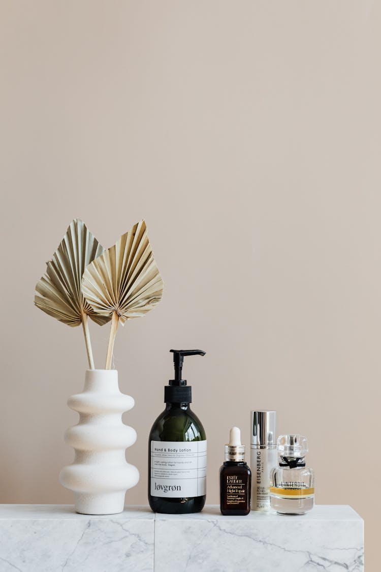 Set Of Cosmetic Supplies Near Decorative Leaves In Vase On Marble Shelf