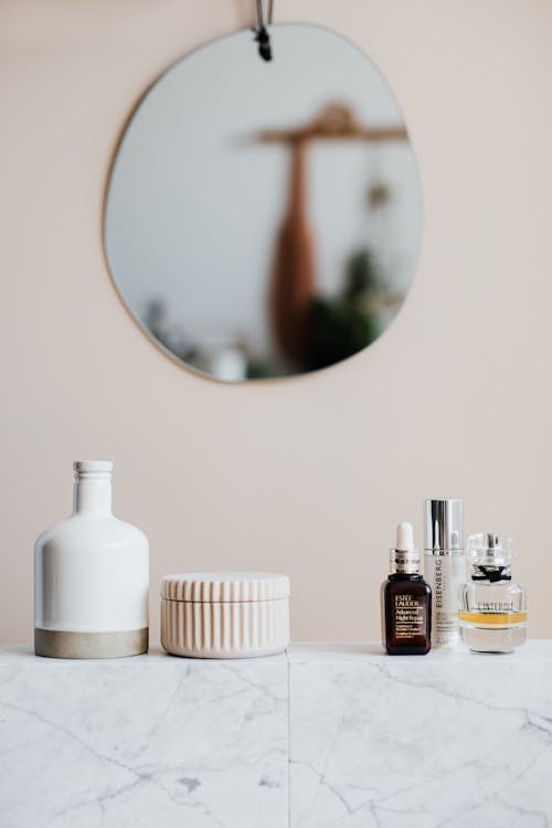 Reusable cosmetic containers for cream and shower gel arranged near various skincare products and perfume bottle on white marble shelf against beige wall with mirror in elegant bathroom interior