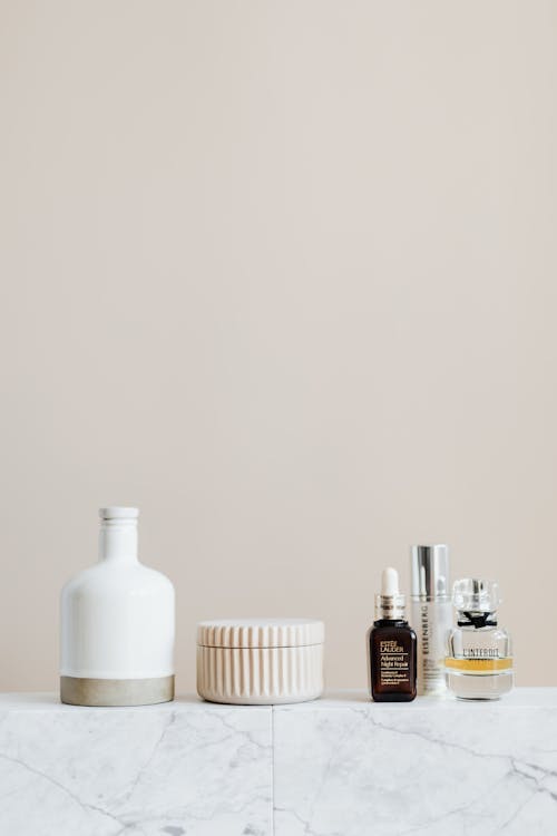 Reusable cosmetic containers for cream and shampoo arranged near various skincare products and perfume bottle on white marble shelf against beige wall in elegant bathroom interior