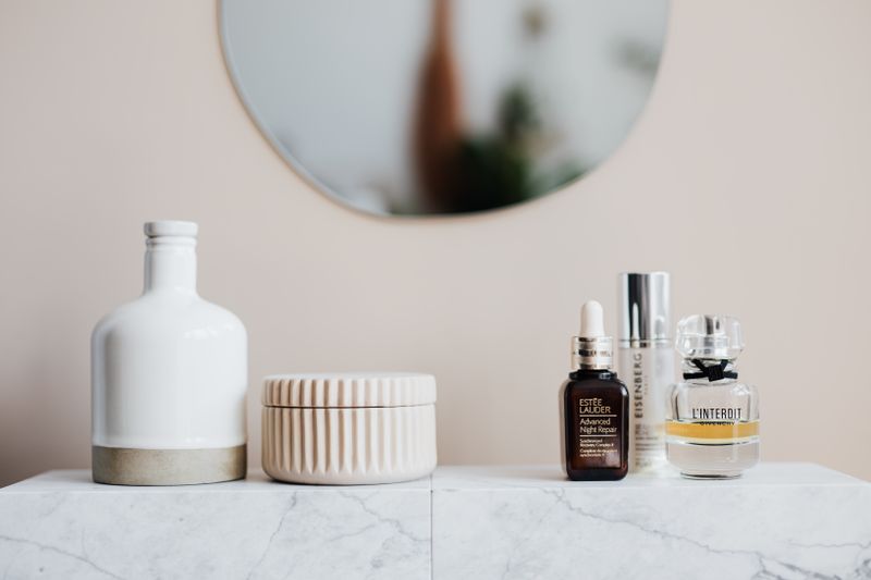 Glass flasks with cream and lotion near perfume and big ceramic vase on marble surface close to beige wall with oval shaped mirror