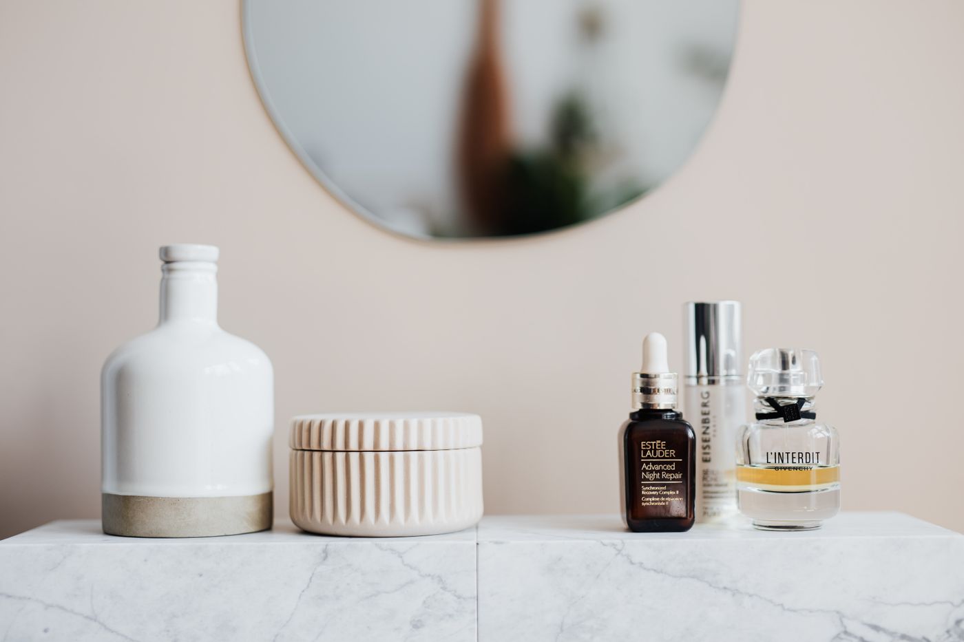 Glass skincare bottles and a ceramic vase on a marble shelf against a warm beige wall