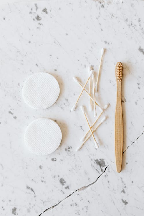 Top view of round cotton pads near pile of buds on thin sticks with soft edges and wooden toothbrush on marble surface