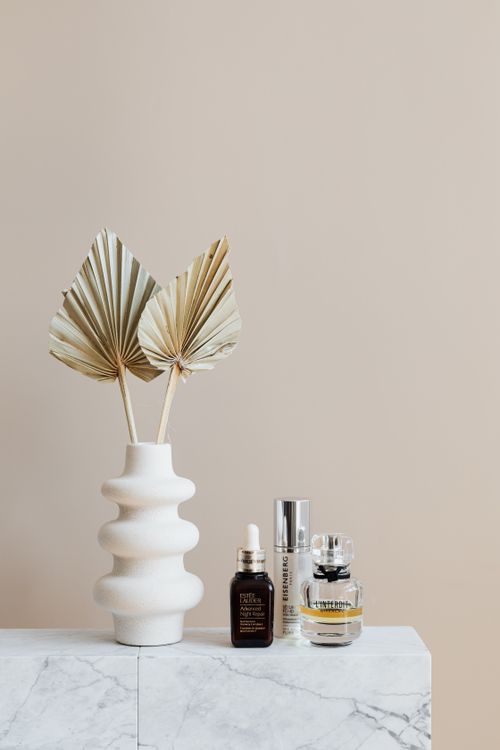 Free Transparent perfume flask near lotions and creative white vase with decorative branches with large ribbed leaves on marble surface near beige wall Stock Photo