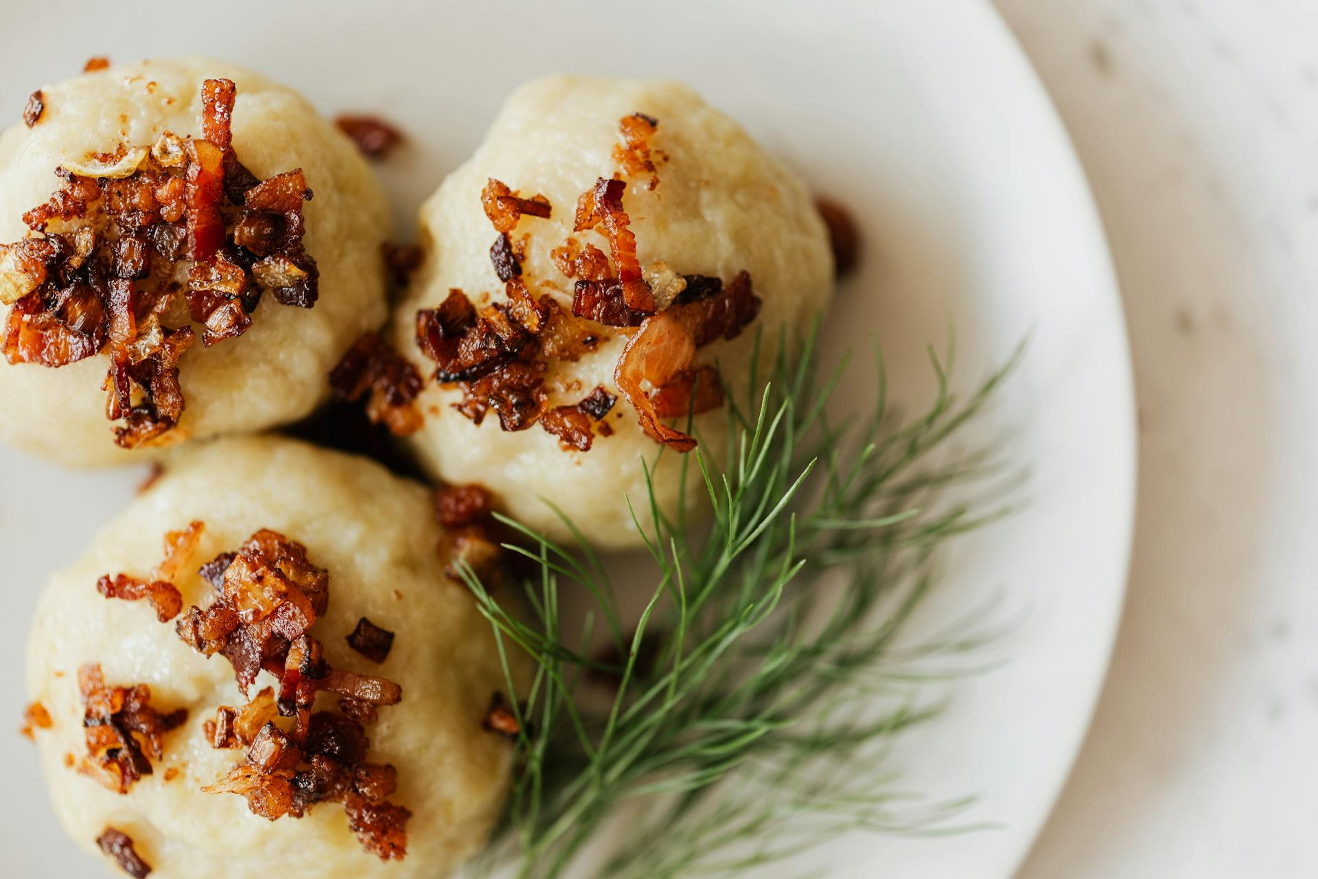 Top view closeup of white ceramic plate with Lithuanian potato cepelinai with fried bacon bits on top and fresh dill sprig aside on marble table