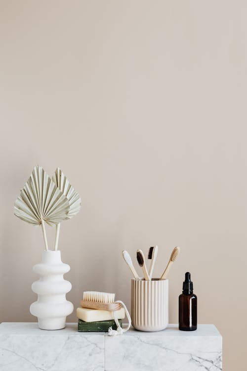 White creative vase with decorative artificial plants and organic soap with body brush placed near set of bamboo toothbrushes in ceramic holder and dark glass reusable bottle of lotion