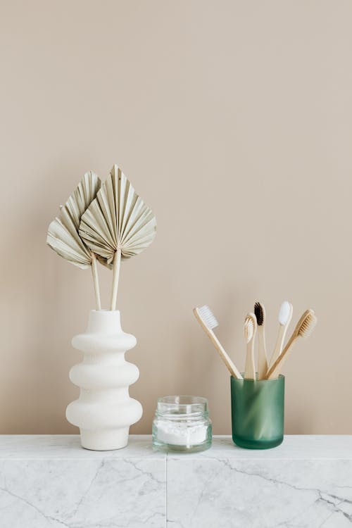Composition of assorted bamboo toothbrushes placed in green holder near organic dental powder in reusable glass jar arranged with creative white vase with artificial flowers