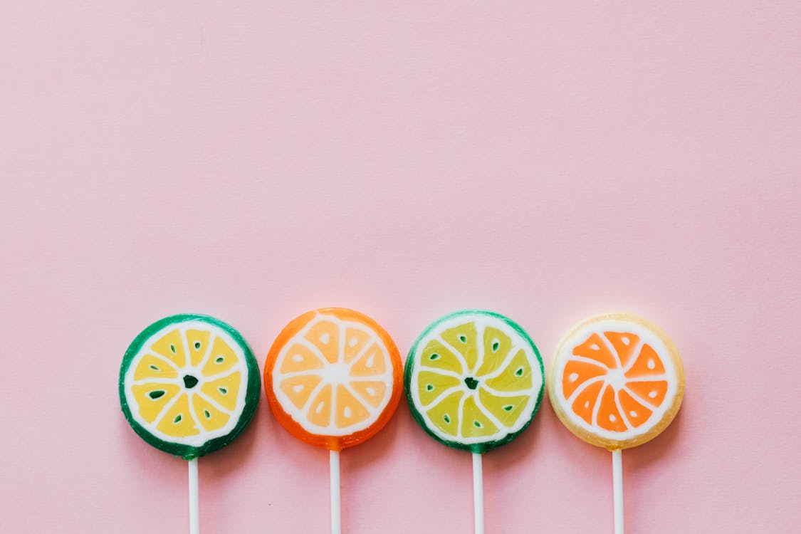 Top view of round multicolored candies with citrus fruit flavor on thin plastic sticks on pale pink surface