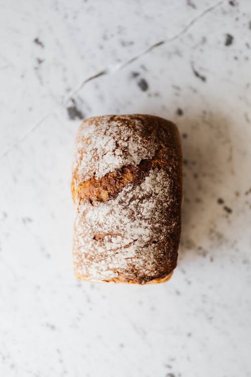 Free Top view of small rectangular baked bread loaf with uneven cracky surface and flour on top on marble table with spots Stock Photo