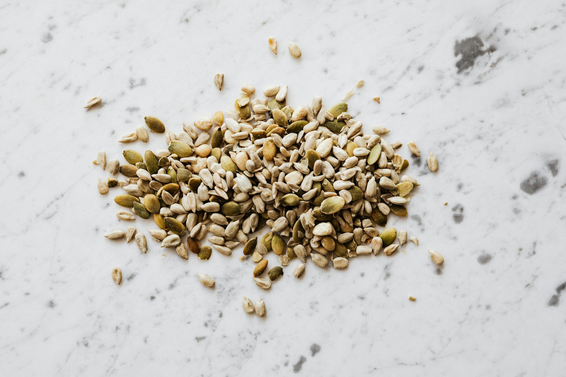 From above of oval shaped dried pumpkin seeds with green and gray shell spilled on marble surface with spots