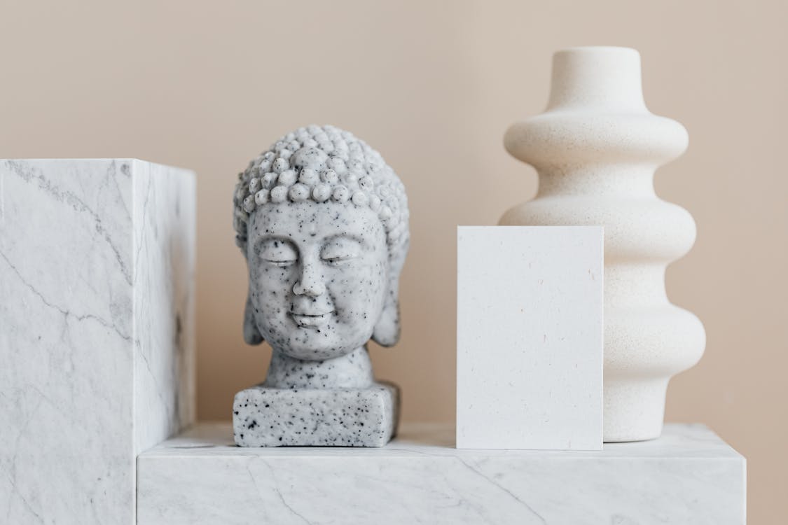 Granite bust of Buddha placed near white ceramic vase of creative geometric shape and blank card on white marble shelf against beige wall