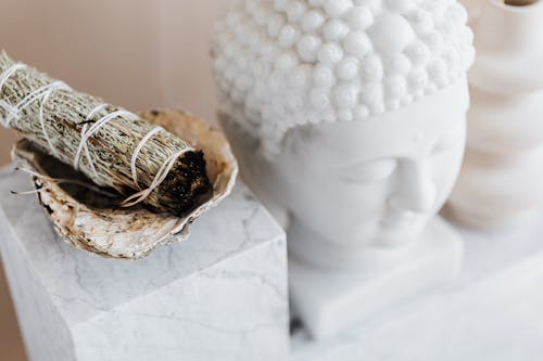 From above of sage candle in bowl placed on white marble shelf near plaster Buddha bust and creative shape vase for home decoration