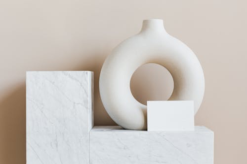 Composition of creative white ceramic vase in ring shape with empty postcard placed on white marble shelf against beige wall as home decoration elements or art objects