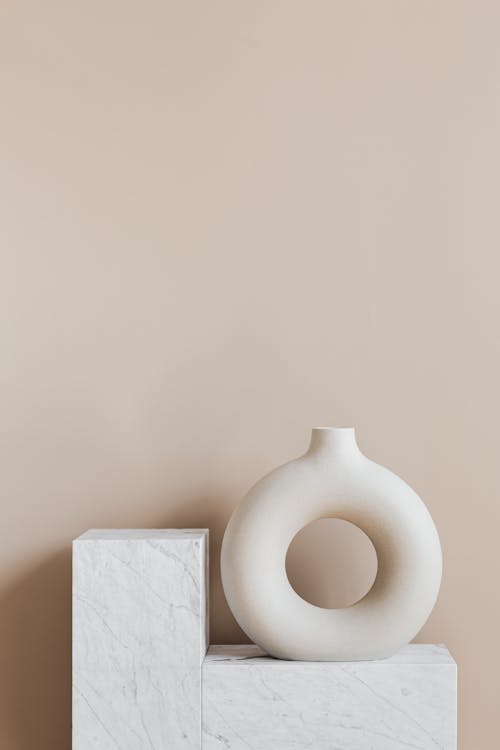 White ceramic vase in creative shape of ring placed on white marble stand against beige wall as home decoration element or art object