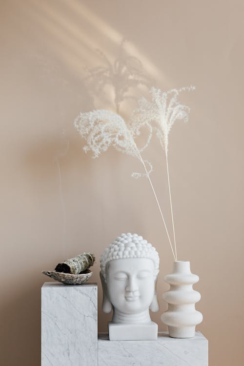 Bowl with sage candle placed on white marble stand near white Buddha bust and geometrically shaped vase with dried herbs for home decoration