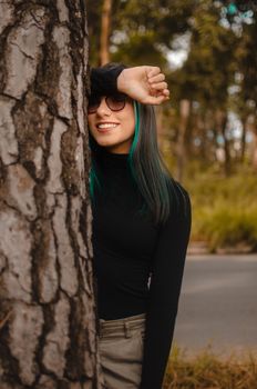 一位开朗的女人，留着绿头发、戴着墨镜，在户外的一棵树旁微笑着
