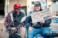 Positive middle aged man showing carton with smile at least your not me inscription while sitting on bench with laughing African American male friend