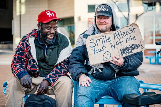 Positive middle aged man showing carton with smile at least your not me inscription while sitting on bench with laughing African American male friend