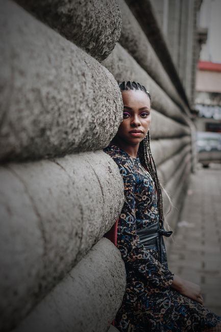 Side view of calm young ethnic lady with Afro braids in casual clothes leaning on wall of aged stone building while resting on street