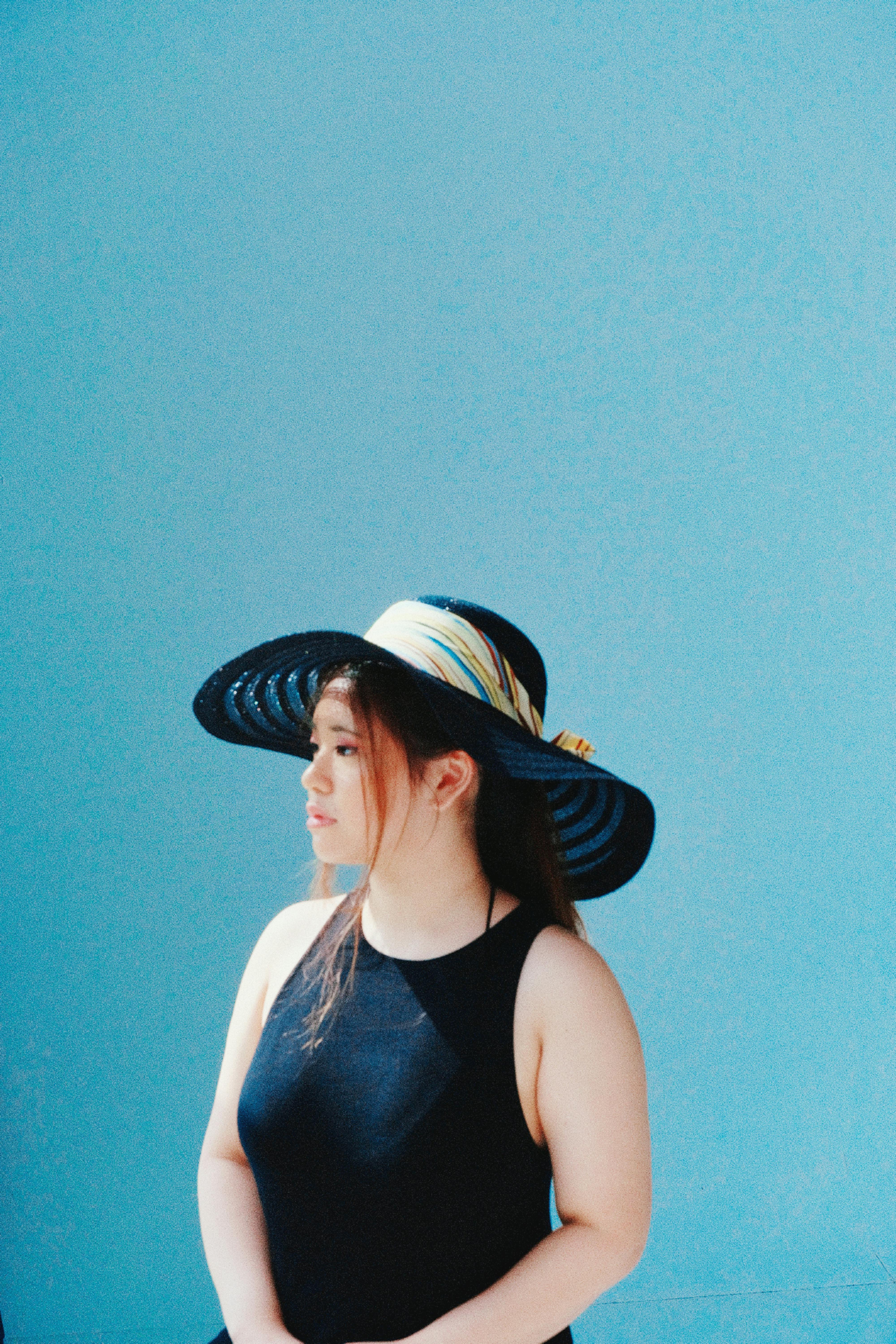 Stylish woman in black top and large sun hat stands against a vivid blue backdrop, creating a striking portrait.