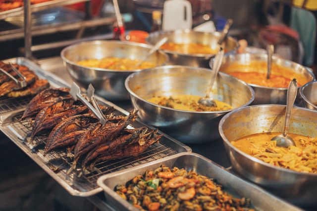 A colorful array of Asian street food dishes in metal bowls on display, showcasing diverse flavors.
